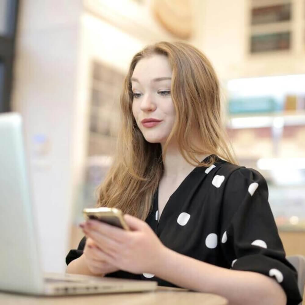 Joven pelirroja utilizando un smartphone y un ordenador portátil en un acogedor espacio de trabajo interior usuario publicando reseñas reales Rating Leader