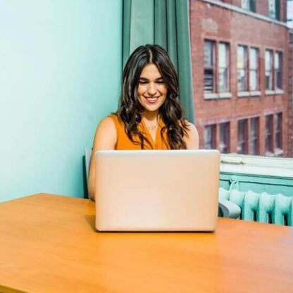 Joven con camiseta naranja empleada de Rating Ledaer trabajando contenta en un ordenador portátil en una mesa de madera en la oficina de Rating Leader junto a la ventana con vistas a un edificio urbano de ladrillo