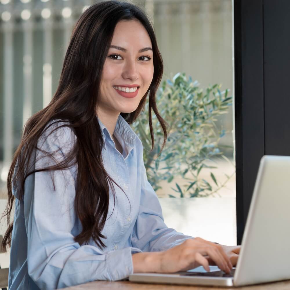 Mujer sonriente con camisa azul trabajando en un ordenador portátil cerca de una ventana con plantas verdes de fondo con el equipo de Rating Leader