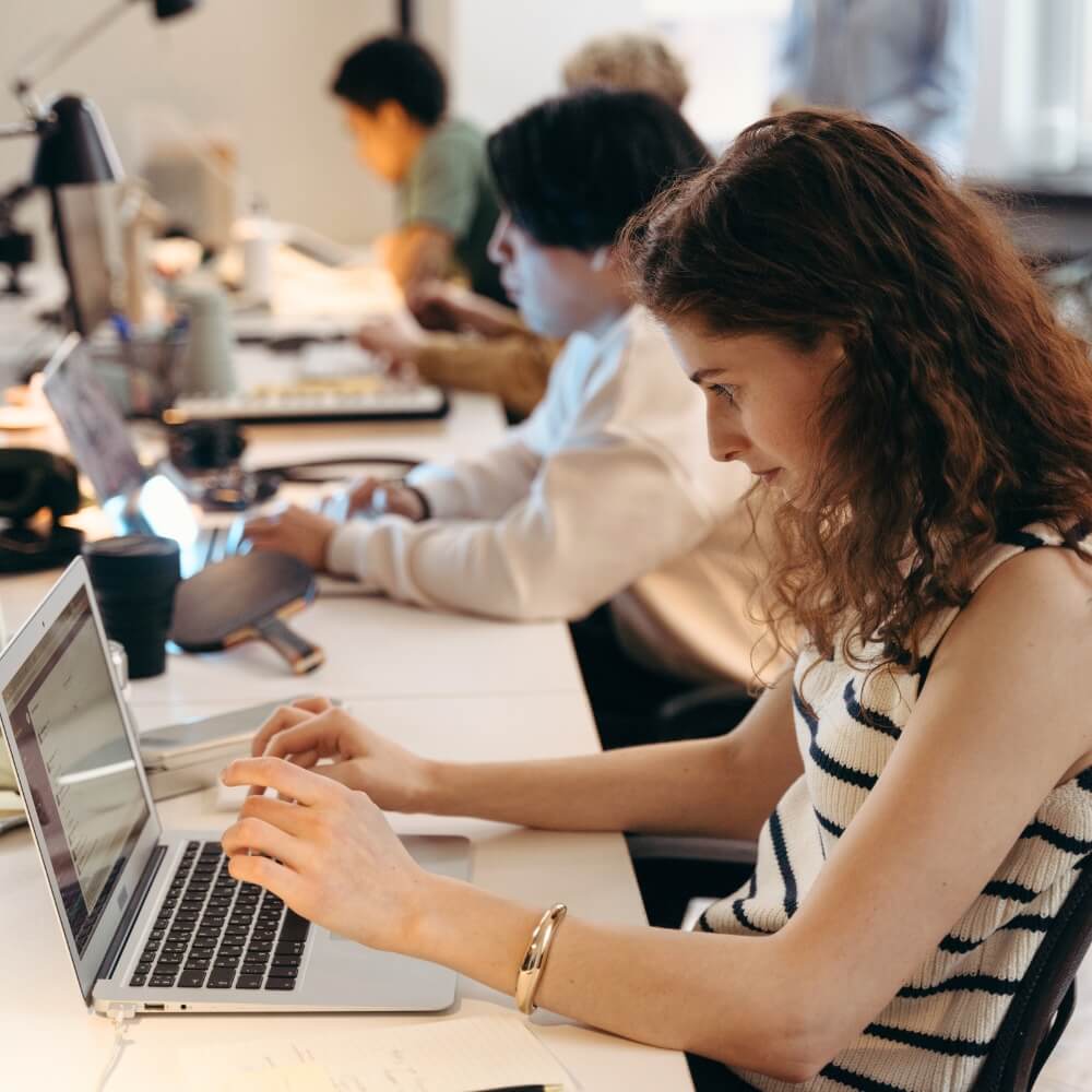 Mujer joven concentrada trabajando en su ordenador portátil en un espacio de trabajo compartido con compañeros al fondo trabajando en servicios de compra de reseñas Google