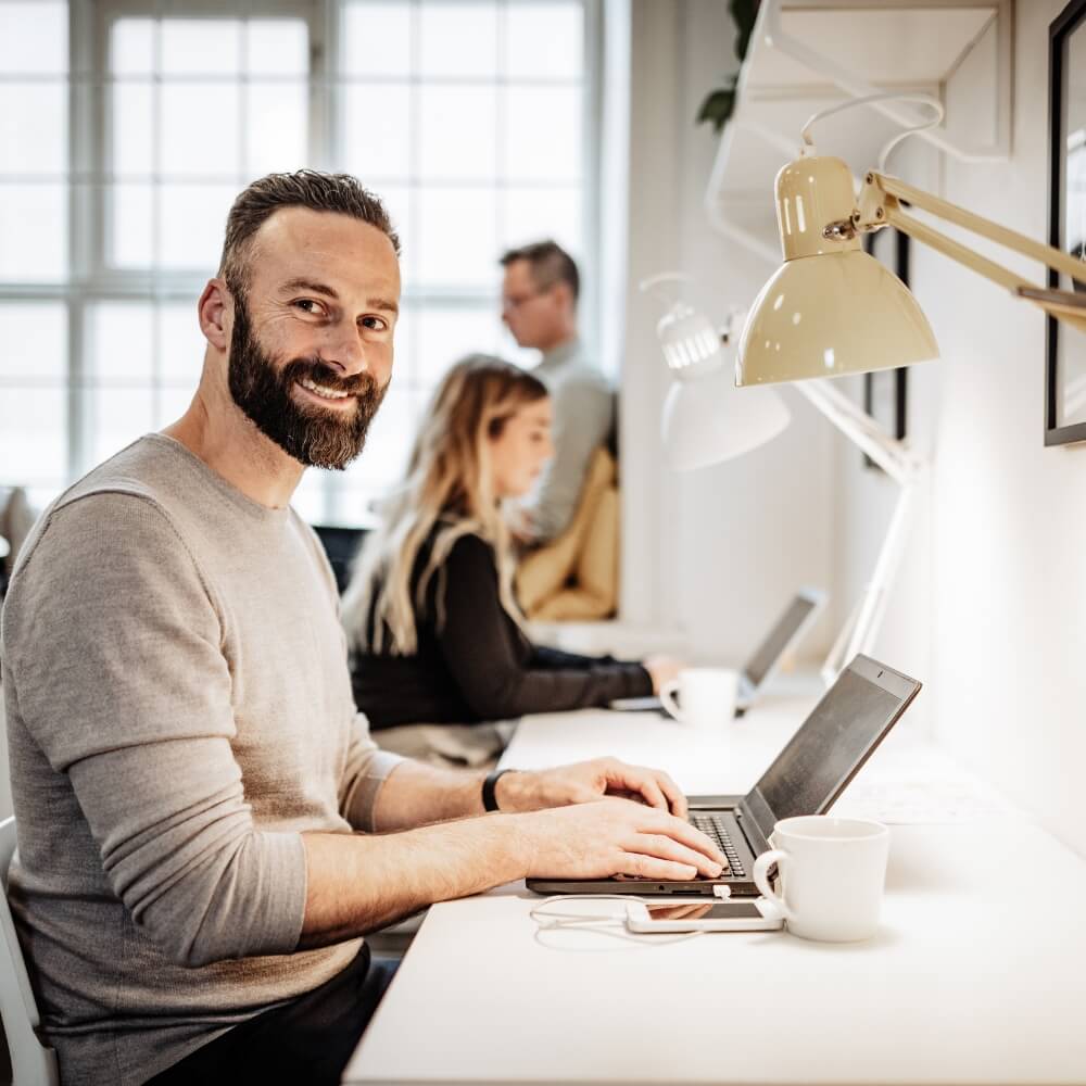 Hombre barbudo sonriente escribiendo reseñas reales para compra de reseñas Google reales en un ordenador portátil en una de las oficinas moderna y luminosas de Rating Leader, compartiendo espacio de trabajo con sus compañeros