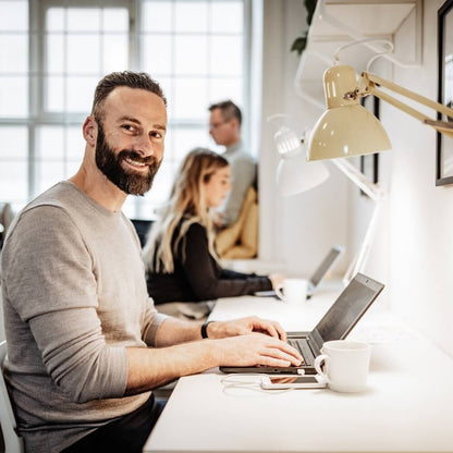 Hombre barbudo sonriente escribiendo reseñas reales para compra de reseñas Google reales en un ordenador portátil en una de las oficinas moderna y luminosas de Rating Leader, compartiendo espacio de trabajo con sus compañeros