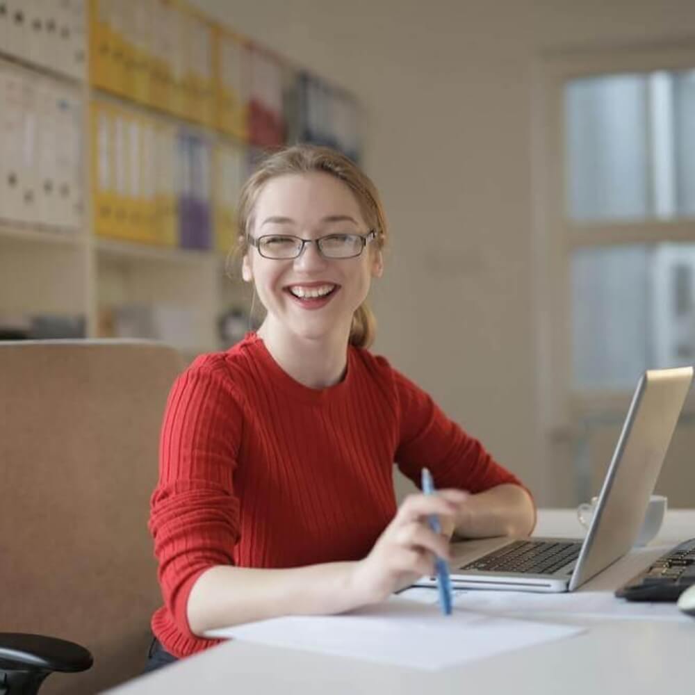 Mujer sonriente con gafas y jersey rojo trabajando con un bolígrafo y un ordenador portátil en una oficina luminosa Rating Leader