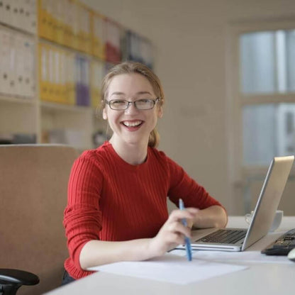Mujer sonriente con gafas y jersey rojo trabajando con un bolígrafo y un ordenador portátil en una oficina luminosa Rating Leader